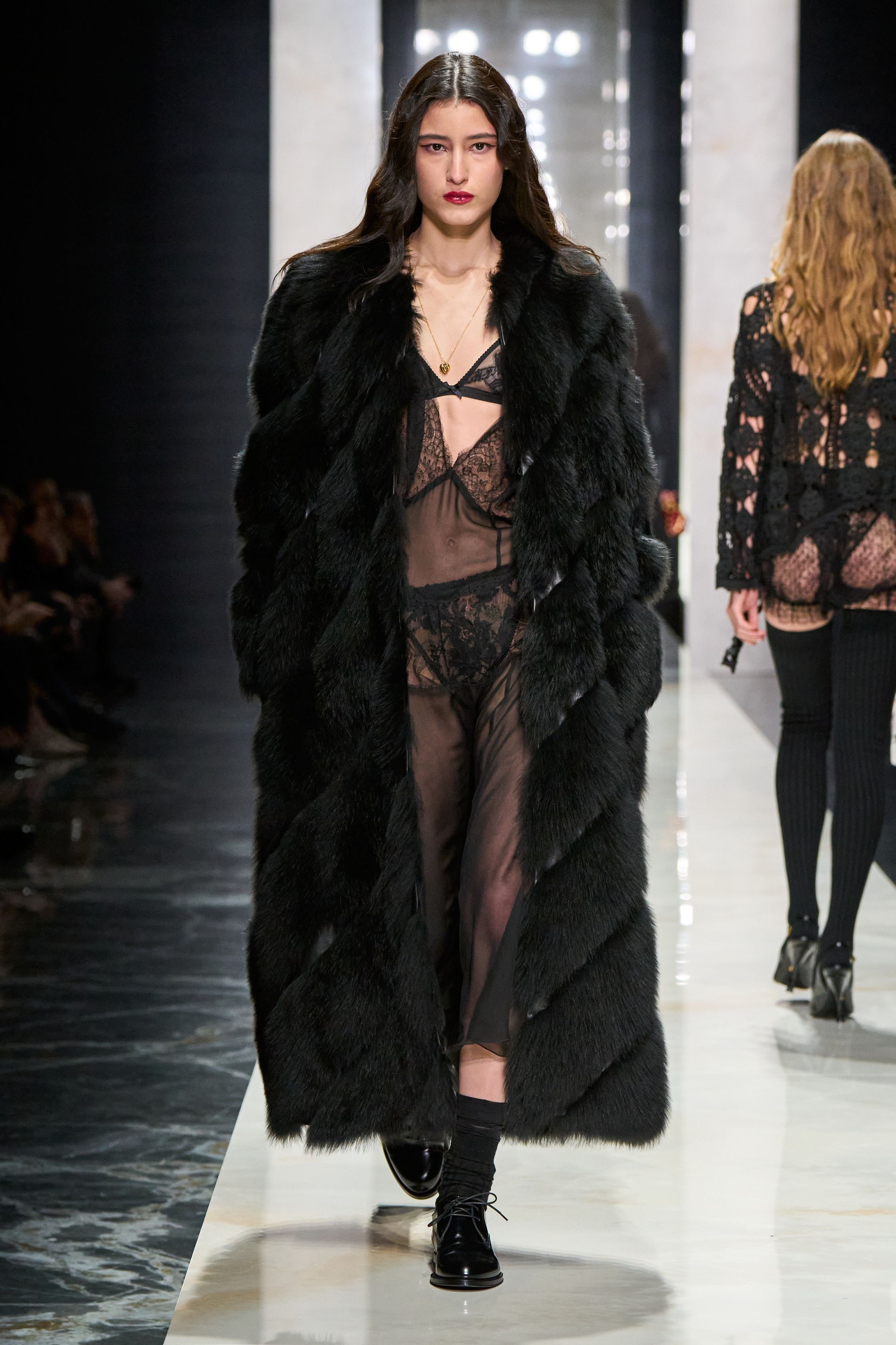Dolce & Gabbana在本季回归品牌标志性的性感意大利美学，通过黑色蕾丝、修身西装与透视礼服塑造极具戏剧张力的女性形象。系列以黑色为核心色调，结合紧身胸衣结构、透明蕾丝裙与宽肩西装，形成刚柔并济的视觉语言。皮草外套与复古帽饰则增添奢华氛围，使整体造型在性感与优雅之间保持平衡，呈现出典型的意大利电影般的华丽气质-顽石频道趋势研究